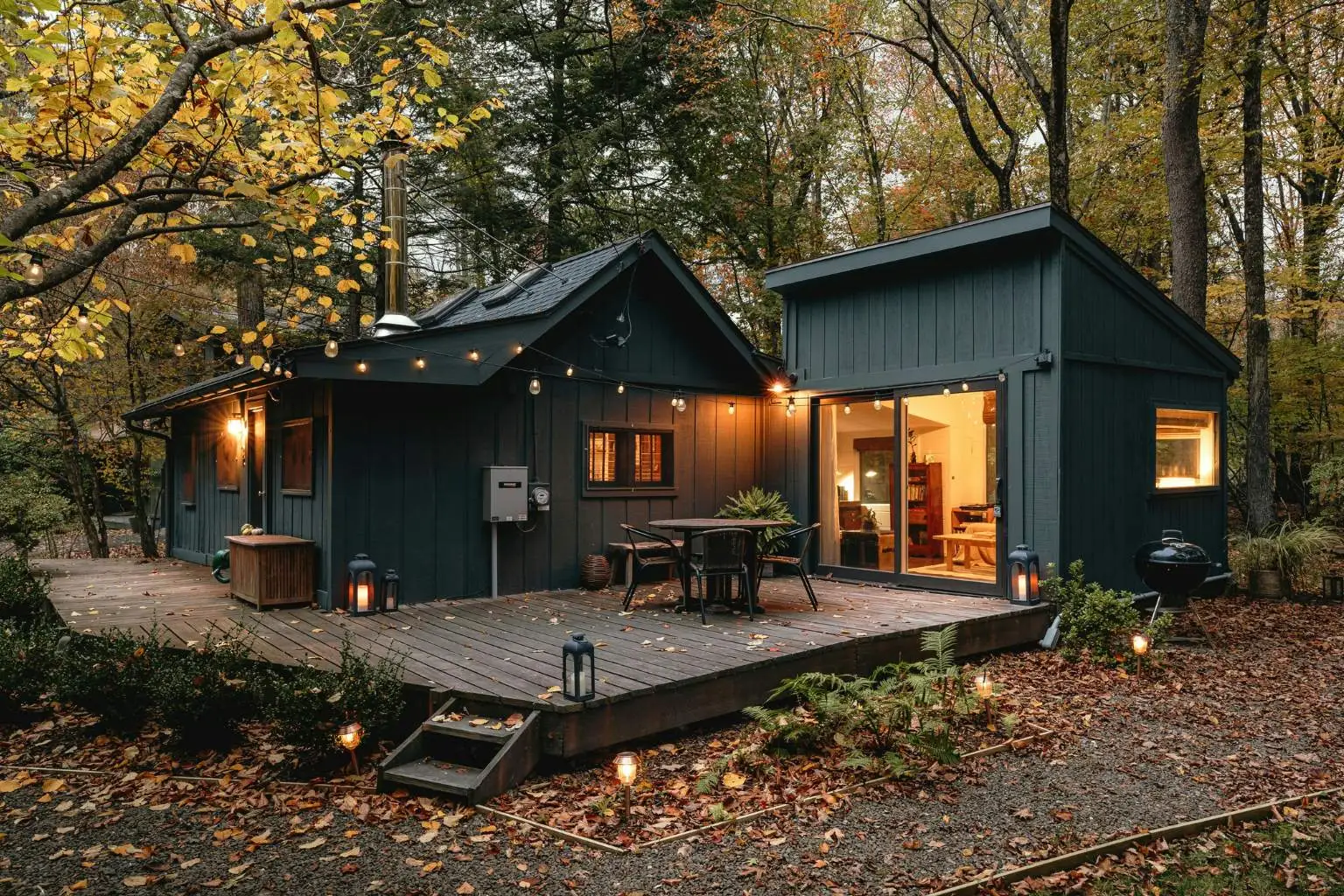 A modern dark gray cabin with a spacious wooden deck nestled in a dense autumn forest, lit up warmly at twilight.